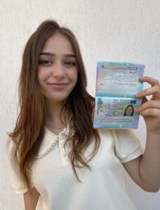 Smiling young woman with long brown hair in a cream sweater holds an open passport toward the camera, wall behind her.